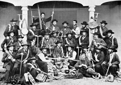 Gruppe von Männern in traditioneller Kleidung mit Wanderstöcken und Ausrüstung vor einem Gebäude.