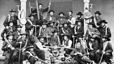 Group of men in traditional clothing with hiking sticks and equipment in front of a building.