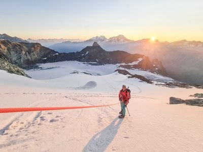 Person mit Helm und Kletterausrüstung auf schneebedecktem Berg bei Sonnenaufgang, Berge im Hintergrund.