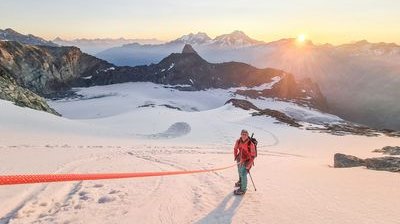 Person mit Helm und Kletterausrüstung auf schneebedecktem Berg bei Sonnenaufgang, Berge im Hintergrund.