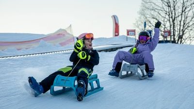 Two people sledding on snow, wearing helmets, goggles, and winter gear, with trees in the background.