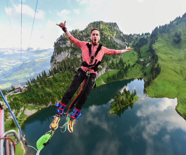Person beim Bungy Jumping über einen See, trägt einen Sicherheitsgurt, Berge im Hintergrund.