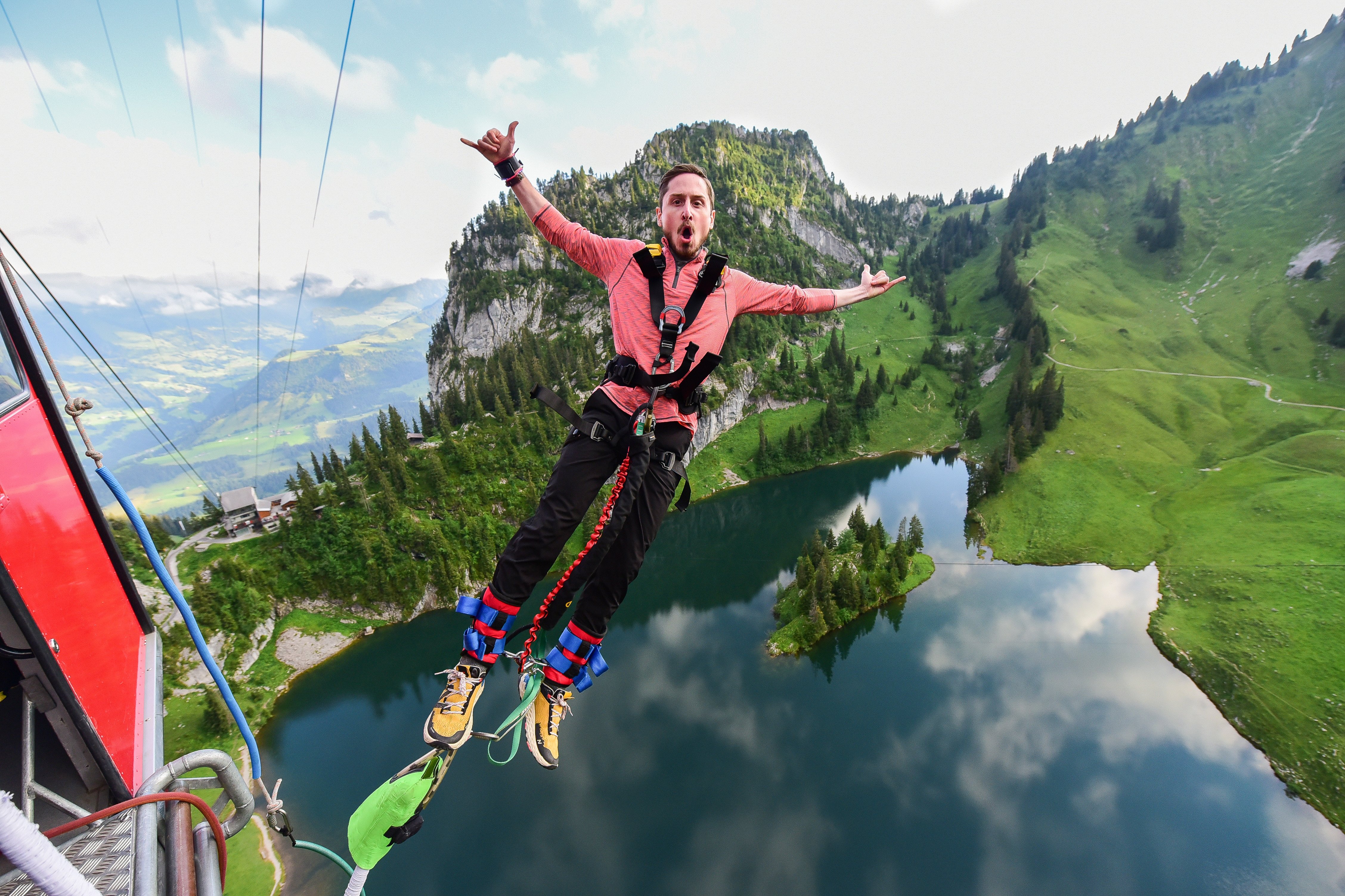Person beim Bungy Jumping über einen See, trägt einen Sicherheitsgurt, Berge im Hintergrund.