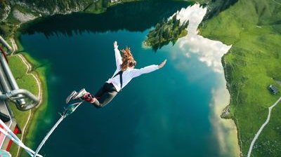 Person beim Bungee-Jumping über einem Bergseeee, trägt ein Sicherheitsgurt und Knöchelriemen, umgeben von Bergen.