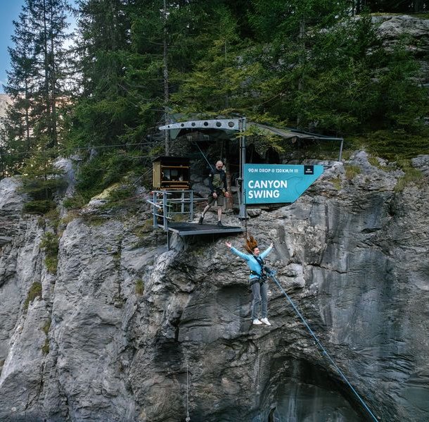 Person in harness jumping from platform for canyon swing, with guide nearby, set in rocky mountain area.