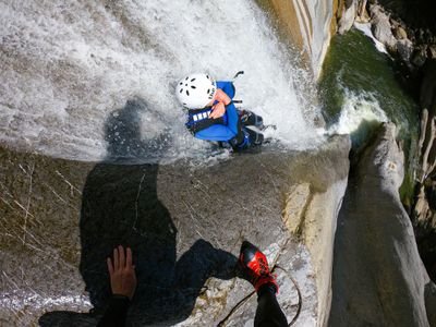 Person in Helm und Neoprenanzug beim Canyoning an einem Wasserfall hinunter, mit sichtbaren Seilen und felsiger Umgebung.
