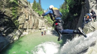 Person in wetsuit and helmet jumps into a canyon pool, others wait on rocks; lush, rocky setting.