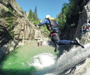 Person im Neoprenanzug und Helm springt in einen Canyon-Pool, andere warten auf den Felsen; üppige, felsige Umgebung.