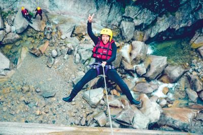 Person in a yellow helmet and red vest rappelling down a rocky canyon, with two others watching.