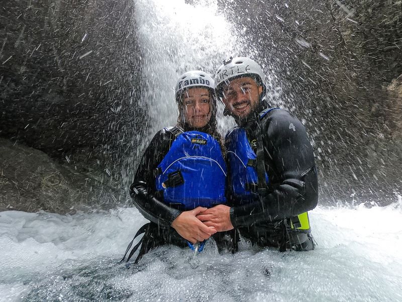 Two people in wetsuits and helmets stand under a waterfall in a rocky canyon, wearing life vests.