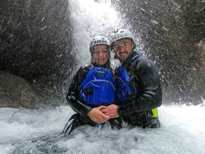 Two people in wetsuits and helmets stand under a waterfall in a rocky canyon, wearing life vests.