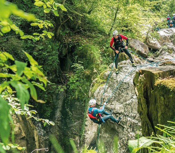 Two people canyoning with helmets and harnesses on a rocky cliff in a lush forest setting.