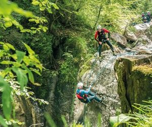 Zwei Personen beim Canyoning mit Helmen und Gurten an einer felsigen Klippe in einer üppigen Waldkulisse.