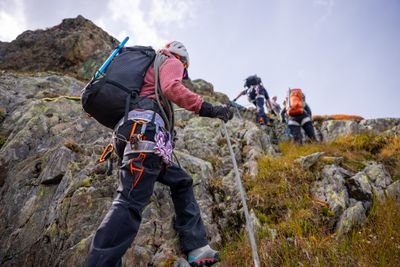 Climbers wearing helmets and harnesses ascending a rocky mountain slope using ropes in daylight.