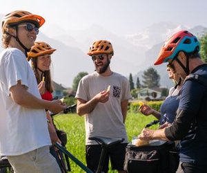 Cyclists wearing helmets stand with bikes in a grassy area, holding snacks, with mountains in the background.