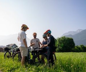 Four people with helmets and bikes stand in a grassy field with mountains in the background.