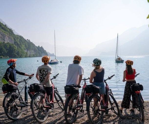 Five people with helmets stand by bikes, overlooking a lake with sailboats and mountain backdrop.