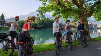 Fünf Personen auf Fahrrädern mit Helmen in der Nähe eines Sees und von Bergen, unter einem großen Baum.