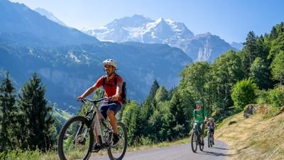 Three people riding e-bikes on a paved mountain road with helmets and backpacks, alpine scenery with snow-capped peaks.