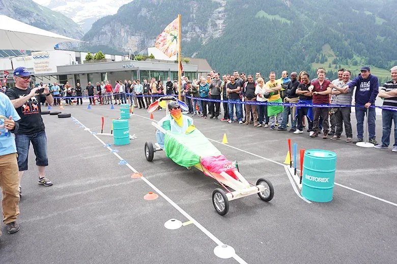 Person in colorful soapbox car on asphalt track, surrounded by spectators in mountain scenery.