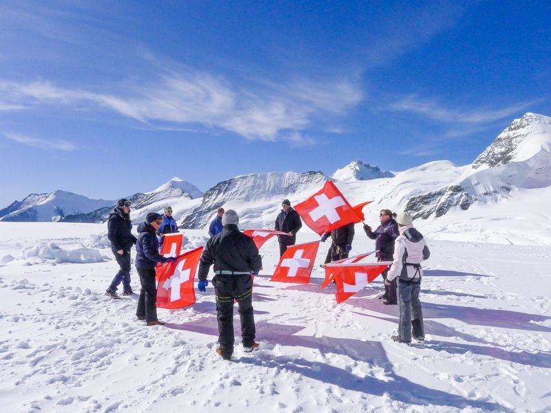 A group with Swiss flags on a snow-covered mountain, some wearing winter clothing and equipment.