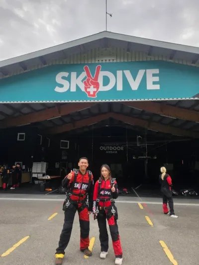 Zwei Personen in Fallschirmsprunganzügen vor einem Gebäude mit "Skydive"-Schild.