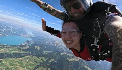 Zwei Personen beim Tandem-Fallschirmsprung über einer Berglandschaft mit See, tragen Helme und Gurte.