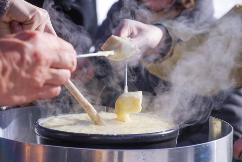 People are dipping bread into a pot of melted cheese outdoors in cool weather.