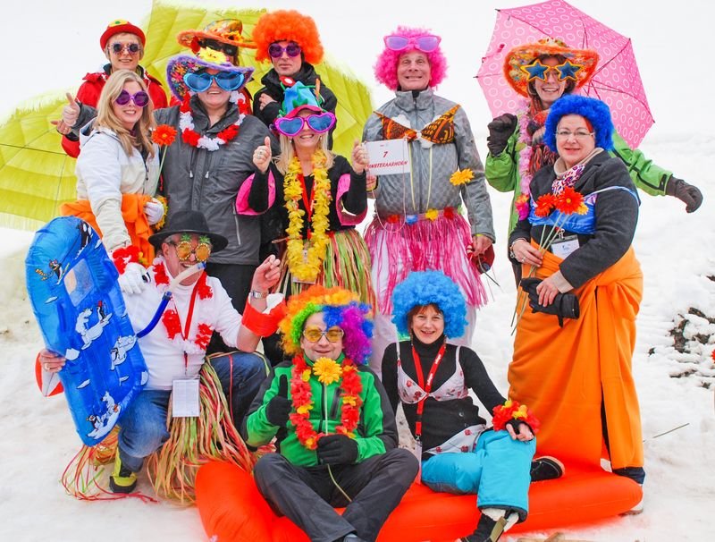 Eine Gruppe in bunten Kostümen und Perücken posiert auf Schnee mit Schlitten und Regenschirmen.
