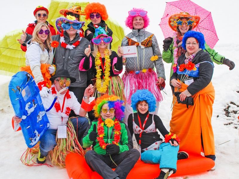 Eine Gruppe in bunten Kostümen und Perücken posiert auf Schnee mit Schlitten und Regenschirmen.