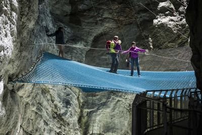 Personen auf einem blauen Netz in einer Felsenschlucht, gesichert mit Seilen, tragen Freizeitkleidung.