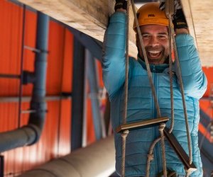 Person in blauer Jacke und Helm klettert drinnen an einer Strickleiter, benutzt Sicherheitsausrüstung und Gurt.