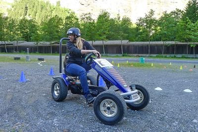 Person on a blue go-kart with a helmet on a gravel surface, surrounded by cones and trees.