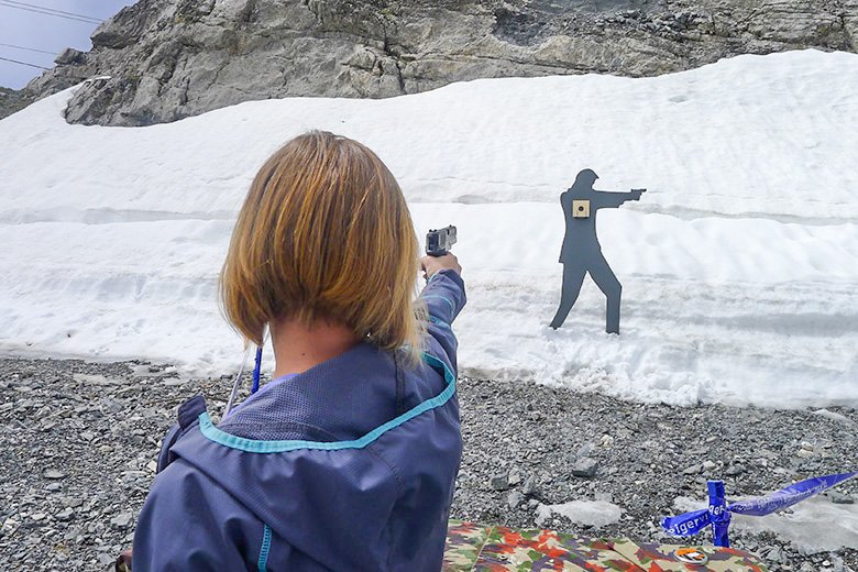Person with jacket aiming a gun at a target in front of a snow-covered rock.