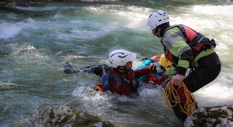 Personen mit Helmen und Neoprenanzügen beim Canyoning in einem Fluss, eine Person mit Seil hilft.