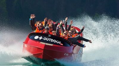 People in a red jet boat with life jackets on a lake, surrounded by splashing water.