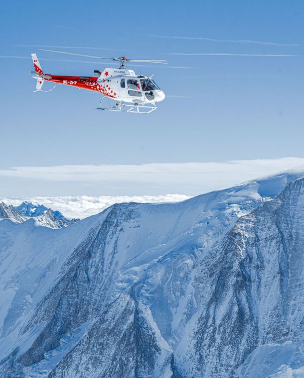 A helicopter flies over snowy mountains under a clear blue sky.