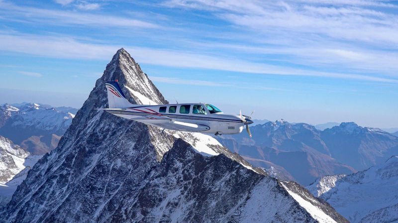 Small airplane flying past a snow-covered mountain peak under a clear blue sky.