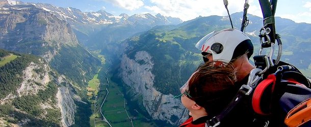 Zwei Personen gleiten mit dem Gleitschirm über ein grünes Tal mit Bergen, sie tragen Helme und Gurte.