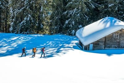 Drei Personen machen Schneeschuhwandern in der Nähe einer schneebedeckten Hütte in einem bewaldeten Gebirgsgebiet und tragen Rucksäcke.