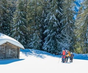 Three people in winter gear snowshoeing in a snowy forest near a wooden cabin.