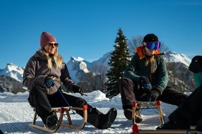 Two people sitting on sleds in snowy mountains, wearing winter clothing and gloves.