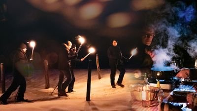 People with torches on a snowy path, next to a table with a steaming pot and bottles.