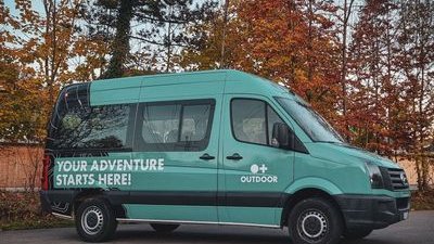 Ein türkisfarbener Van mit der Aufschrift "Your Adventure Starts Here!" steht vor herbstlichen Bäumen.