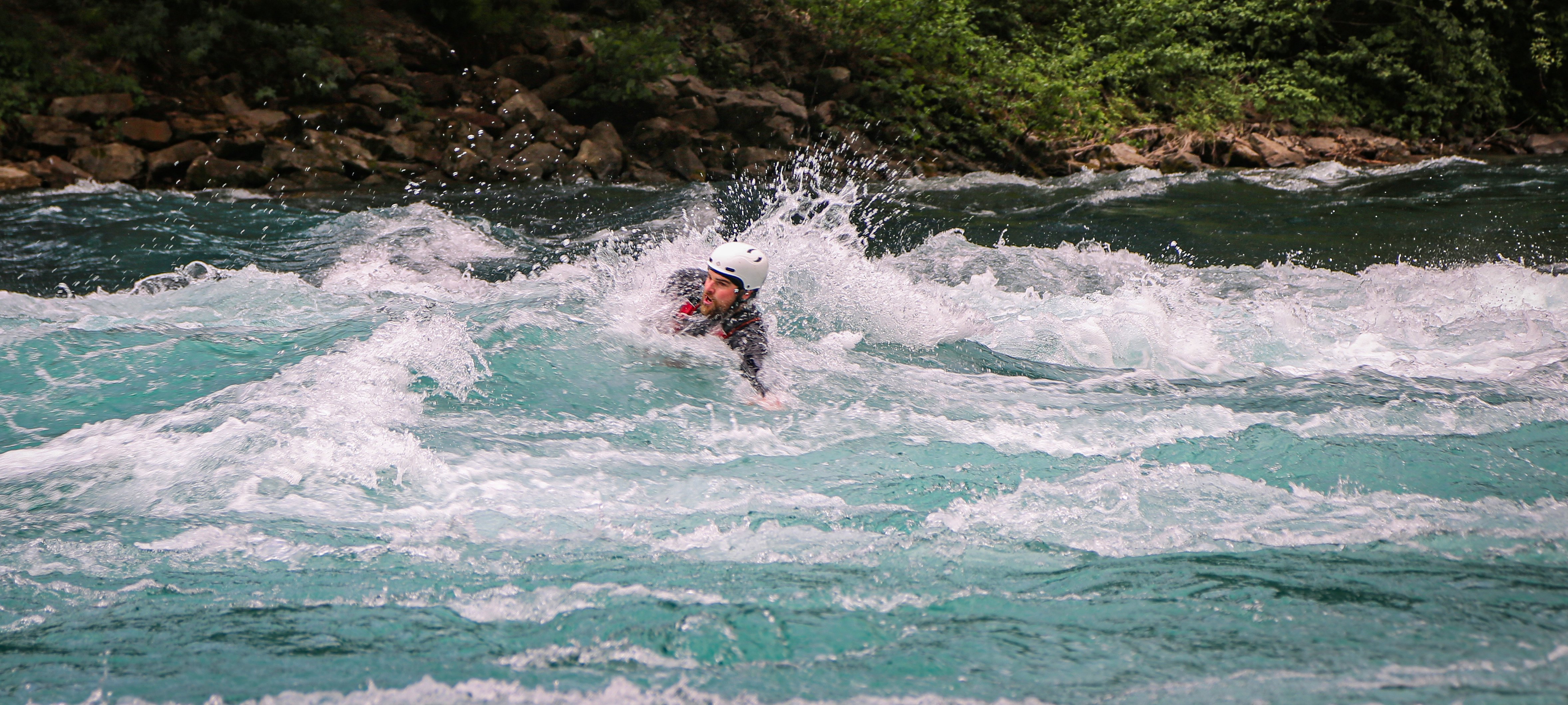 Person im Neoprenanzug und Helm schwimmt im Wildwasserfluss, umgeben von Bäumen und Felsen.