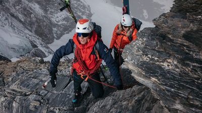 Two people are climbing a rocky mountain with helmets and ropes, skis attached to their backpacks.
