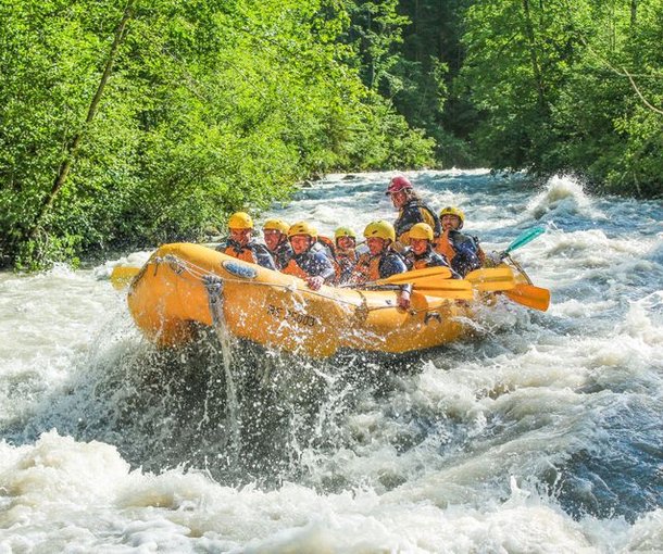 Gruppe in gelben Helmen und Rettungswesten beim Wildwasser-Rafting auf der Simme, umgeben von Bäumen.