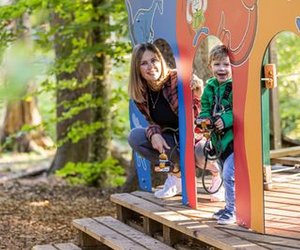 Child with safety harness, accompanied by adult on a wooden platform in the Interlaken Rope Park in the forest.