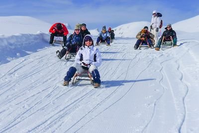 People are sledding on a snowy slope in the mountains, wearing winter clothes and helmets.