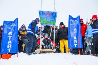 People in winter clothing and helmets start a sled race in the snow between blue banners.